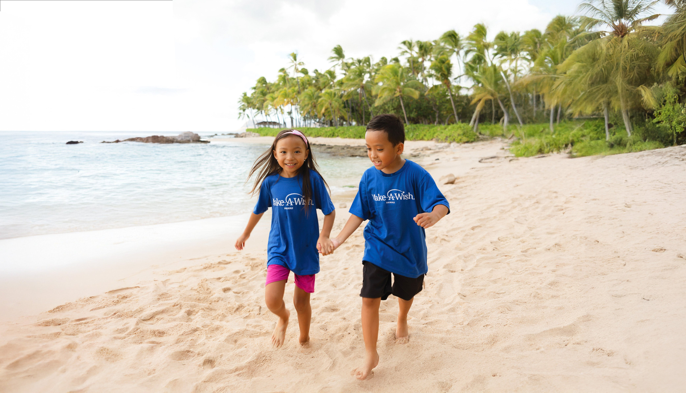 Two children in Make-A-Wish Hawaii shirts walk hand in hand on a beach.
